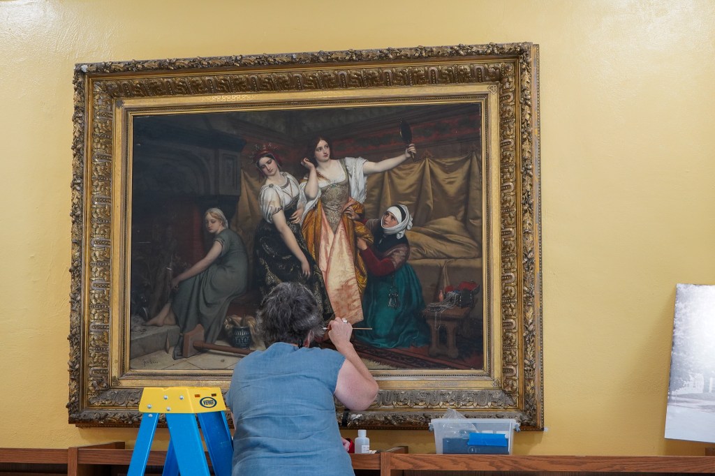 A photo of a woman cleaning an oil painting with a small q-tip, the painting has a large gold frame and hanging on the library wall.